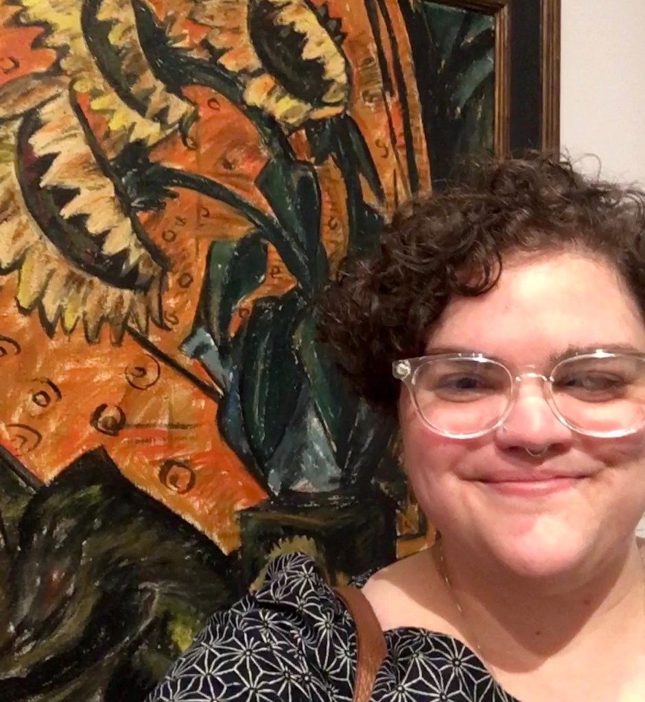 Hannah taking a selfie in front of an oil painting depicting sunflowers. She is a white woman with short, brown, curly hair, and clear plastic-framed glasses.